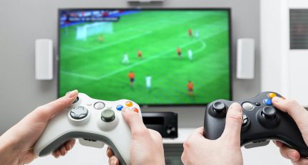 Two white game controllers held by hands in front of a TV screen displaying a virtual soccer match