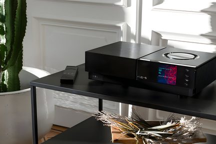 Amplificateur audio noir avec écran LED bleu sur une table en bois, à côté d'un vase avec des branches séchées