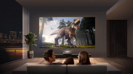 Salon moderne avec écran géant affichant un dinosaure, 3 personnes de dos sur un canapé gris, plante verte en pot