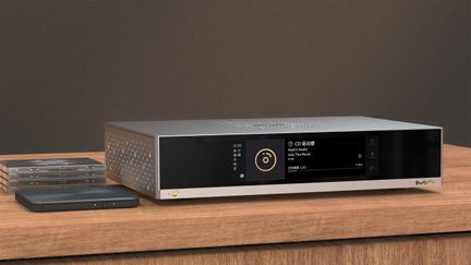 Προβολή τριών τετάρτων από μπροστά ενός ορθογώνιου audio boîtier από ασημί αλουμίνιο, με μαύρη γυάλινη πρόσοψη που ενσωματώνει έγχρωμη οθόνη, μικρό στρογγυλό χρυσό κουμπί κάτω αριστερά, χρυσό λογότυπο δεξιά και πλαϊνή πλευρά με διατρητά μοτίβα ρόμβου, τοποθετημένο πάνω σε ξύλινη βάση.