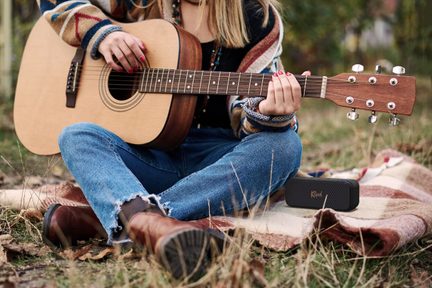 Kobieta siedząca na trawie gra na drewnianej gitarze akustycznej, ubrana w podarte dżinsy i wełniany sweter w wzory, brązowe buty