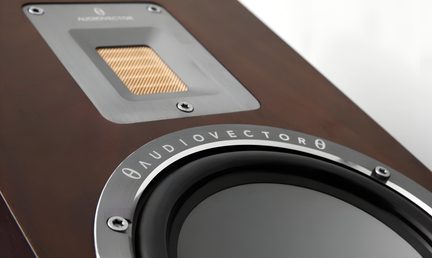 Gros plan oblique sur la façade en bois brun d’une enceinte, montrant un tweeter ruban à grille dorée encadré d’aluminium brossé et un woofer noir cerclé d’un anneau métallique gravé « AUDIOVECTOR »