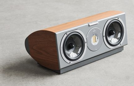 Enceinte centrale finition bois brun et façade métallique grise, deux haut-parleurs circulaires à membrane tissée encadrant un tweeter ruban vertical, boîtier aux flancs arrondis, vue de trois-quarts avant gauche