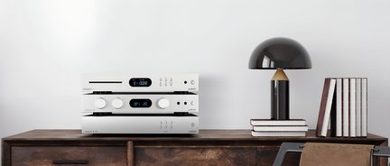 Trois appareils audio empilés avec écrans noirs et boutons argentés, lampe noire en dôme, livres empilés sur un meuble en bois brun