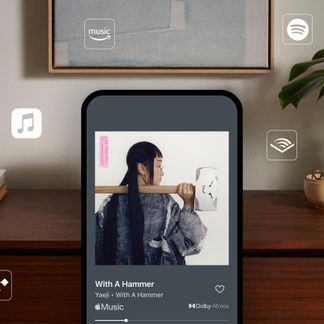 Écran intelligent rectangulaire noir mat en plastique, vu de face, affichant une pochette d’album, posé sur un plateau en bois sombre, entouré d’icônes de services audio flottantes, avec cadre décoratif et plante verte en arrière-plan