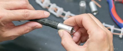 Mains tenant un câble gainé noir avec connecteur métallique cylindrique argenté, arrière-plan flou avec outils et composants