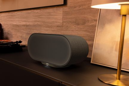 Diffusore wireless Denon nero a forma ovale, con griglia sottile, posizionato su un mobile TV in un soggiorno moderno con illuminazione calda