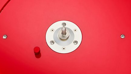 Gros plan vue de dessus d’une surface rouge laquée avec plaque circulaire en métal argent fixée par quatre vis abritant un commutateur cylindrique satiné, accompagné d’un petit bouton moleté rouge à gauche