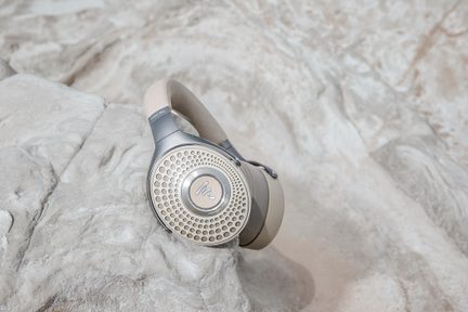 Beige och gråmetallic circumaurala hörlurar med perforerad metallram på kåporna och vadderat huvudband i läderlook, visade i trekvarts vy placerade på en stenig yta