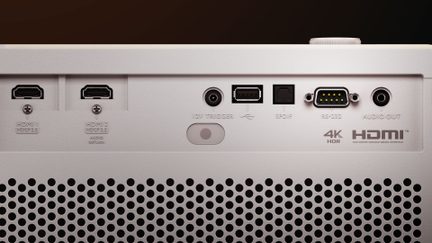 Bakifrån i närbild av en vit matt plastvideoprojektor med två HDMI-portar, en USB-port, en optisk SPDIF-anslutning, ett seriellt RS-232-uttag, 12 V-ljuduttag och 3,5 mm jackuttag, samt ett stort perforerat ventilationsgaller.
