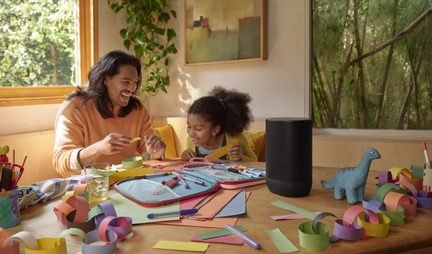 Haut-parleur cylindrique noir en plastique à grille perforée intégrale avec base plus épaisse, vue de trois-quarts avant, posé sur une table en bois parmi fournitures de loisirs créatifs