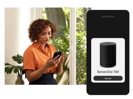Ženska s kodrastimi lasmi drži črn pametni telefon v roki in gleda na zaslon, na desni strani je povezani zvočnik Sonos Era 100