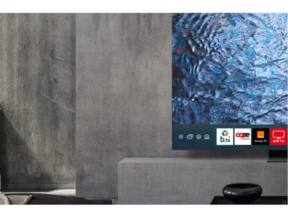 Téléviseur Samsung TQ65S95D 2024 affichant une image abstraite bleue, posé sur un meuble gris dans une pièce en béton
