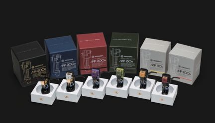Six cellules phono sur porte-cellules tubulaires noirs, blocs frontaux métalliques noir, argenté, violet, vert, orange et rouge, alignées sur socles blancs en mousse rigide avec câbles bleus visibles, six boîtes cartonnées assorties à l’arrière, vue frontale légèrement en plongée