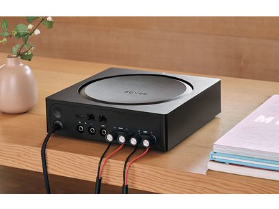 Črn stereo hi-fi ojačevalnik Sonos Amp, pogled od spredaj, priključki na zadnji strani, vrtljivi gumb na vrhu, postavljen na leseno mizo