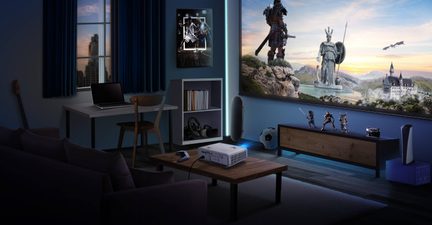 Sned vy av ett multimedierum med blågrå väggar, där en vit plastbordprojektor står på ett lågt träbord och projicerar en stor fantasyscen på en väggmonterad projektionsduk, omgiven av en lila soffa, ett ljusgrått skrivbord med bärbar dator, en låg träförvaring och blå LED-belysning