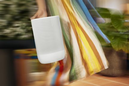 Enceinte cylindrique blanche en plastique mat et grille métallique perforée, logo vertical Sonos, vue de trois quarts avant, tenue par une main devant un arrière-plan flou