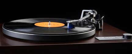Platine vinyle vue de trois-quarts avant, base en bois brun verni, plateau noir portant un disque à étiquette orange, bras gris métallique terminé par une cellule bleue et molette de réglage noire sur le coin droit