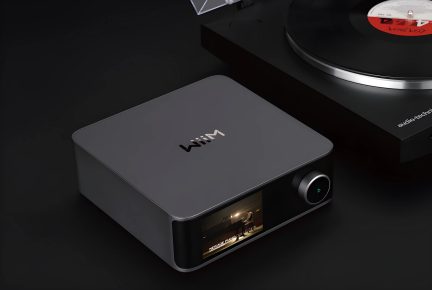 Boîtier rectangulaire noir avec écran en façade, bouton circulaire à droite, platine vinyle partiellement visible à l'arrière