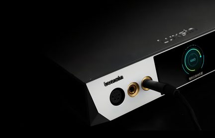 Primer plano en ligera vista cenital de la fachada frontal plateada de una caja de audio rectangular negra de aluminio, equipada con una pantalla circular retroiluminada y tres conectores para auriculares con anillos dorados, uno de los cuales está ocupado por un cable negro.