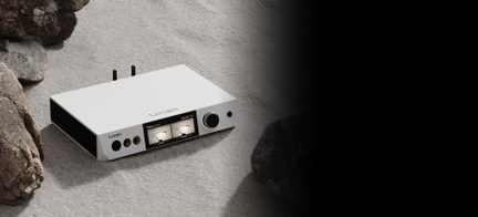 Caja rectangular de aluminio blanco, vista en tres cuartos desde arriba, panel frontal con pantalla central de doble vúmetro, gran botón negro a la derecha, dos tomas de auriculares doradas a la izquierda, laterales negros con aletas de disipación, dos antenas negras en la parte trasera