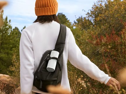 Enceinte portative cylindrique gris clair Sonos à grille métallique perforée, fixée sur le côté d’un sac à dos noir vue arrière