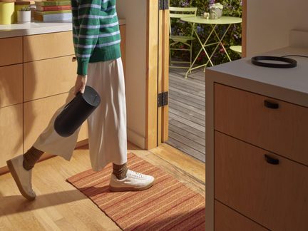 Vue latérale d’une enceinte portable cylindrique noire en plastique mat avec surface supérieure à commandes tactiles, tenue à la main par une personne marchant sur un sol en bois dans une cuisine
