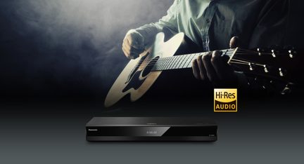 Melns audio atskaņotājs no matēta metāla un spīdošā plastmasas, skats no priekšas, gluda priekšpuse ar centrālo digitālo ekrānu, kas rāda 0:00.00, un balto logo pa kreisi.