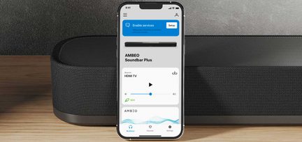 Pametni telefon z vklopljenim zaslonom, ki prikazuje vmesnik aplikacije AMBEO Soundbar Plus, postavljen na črno zvočno letev, svetla lesena površina