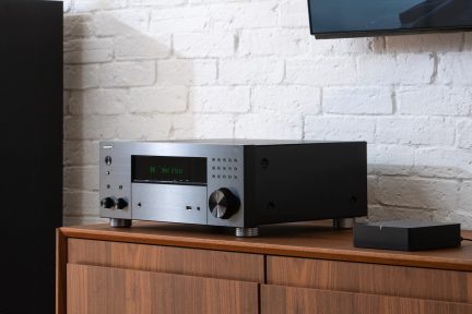 Black and silver amplifier, circular knobs, green digital display, placed on wooden furniture, white brick wall