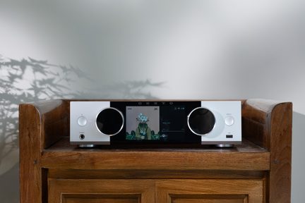 Srebrny, podłączony wzmacniacz Hi‑Fi Onkyo z dużym kolorowym ekranem pośrodku i dwoma szerokimi, okrągłymi pokrętłami, ustawiony na drewnianej szafce