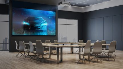 Salle de réunion avec table rectangulaire en bois, 12 chaises grises, écran mural affichant une voiture, sol en bois, fenêtres vitrées