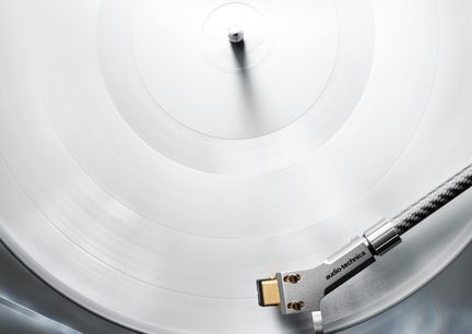 Lähivaade ülalt valgele vinüülplaadile, mis asub plaadimängijal; hõbedane toonarm on valmistatud metallist ja süsinikkiust, kuldne helipea kannab « audio-technica » logo.
