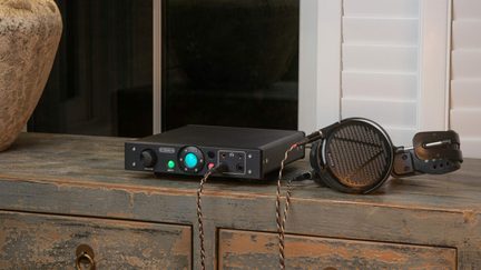 Črni kovinski namizni audio ojačevalnik in priključen odprti okoloušesni slušalnik, pogled od sprednjega tričetrtinskega kota, z prikazanim gumbom za glasnost, osrednjim retro osvetljenim zelenim krogom, dvema priključkoma za slušalke in priključenim pletenim kablom, postavljenim na leseno podlogo