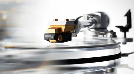 Złota metalowa wkładka gramofonowa z czarną plastikową obudową, widok z profilu w dużym zbliżeniu, zamocowana na srebrnym ramieniu nad przezroczystą płytą winylową