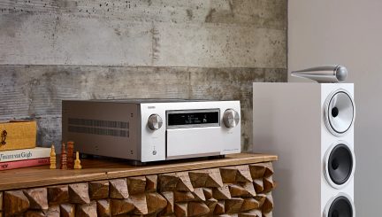 Amplificateur argenté avec boutons circulaires, écran central, posé sur meuble en bois sculpté, enceinte blanche à droite