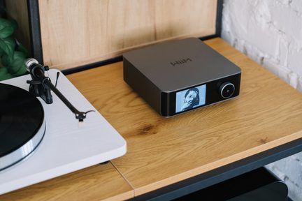 Chaîne hi-fi composée d’un boîtier audio gris anthracite en métal, vue de trois-quarts avant légèrement plongeante, avec façade noire brillante intégrant écran couleur et molette circulaire, posé à droite d’une platine vinyle blanche au plateau noir sur une étagère en bois