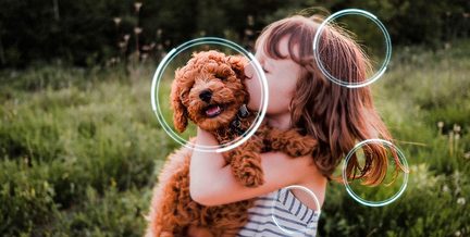 Fillette brune en robe rayée en tissu blanc et bleu vue de face serrant un chiot bouclé roux au collier noir dans une prairie verdoyante avec quatre cercles lumineux bleutés superposés