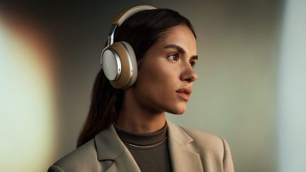 Trådlösa over-ear-hörlurar i tvåfärgad matt guld och silver, metallkåpor med ljusbeige öronkuddar i konstläder, vadderat huvudband och synlig logotyp, vänster sidovy på en persons huvud