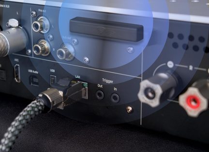 Lähiplaanis musta mati metallist heliseadme tagakülg: ristkülikukujuline must wifi-piktogrammiga dongel, RCA pre-out ja sub-out pesad, USB-port, LAN-portis punutud kaabel, mini-jack trigger-pesad ning punased ja hallid kõlariterminalid.