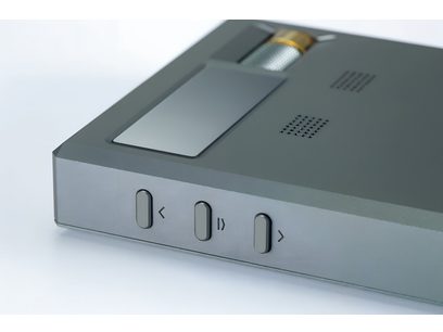 Nära vänster framifrån-vy av den bärbara ljudomvandlaren Cayin RU9 i matt grå aluminium, med tre ovala svarta knappar märkta <, paus/uppspelning och > på kanten, en ovansida med ett rektangulärt glasfönster, en silverfärgad gallercylinder med guldring och två perforerade ventilationsgaller.