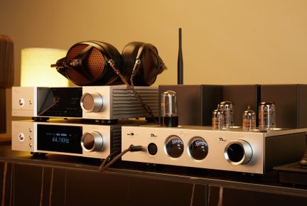 Silver brushed metal hi-fi set seen from a three-quarter front angle, featuring a tube amplifier with three circular VU meters and a large volume knob, two stacked digital converters with blue displays and potentiometers, topped with a black over-ear headphone with copper grills