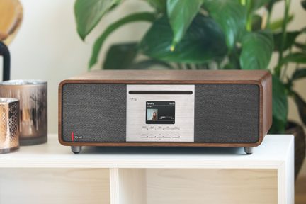 Chaîne audio compacte rectangulaire en bois brun avec façade frontale combinant panneau central en métal brossé argenté intégrant écran couleur, fente et rangée de boutons, entouré de grilles de haut-parleur en tissu gris foncé, vue de face