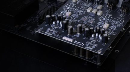 Mustast klaaskiust trükkplaat, pealtvaates kaldnurgast, varustatud mustade ja hõbedaste silindriliste elektrolüütkondensaatorite, pinnale paigaldatud integraallülituste, lameda pistiku ja metallist vahetükkidega