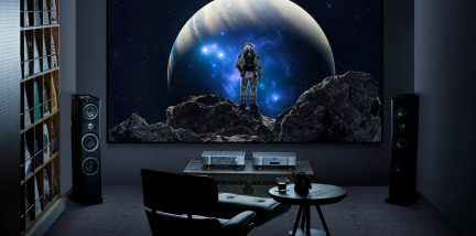 schermo rettangolare di grande formato, immagine di astronauta su sfondo spaziale, due diffusori a colonna neri ai lati, mobile elettronico basso, poltrona nera, tavolino rotondo, vista frontale di una stanza buia