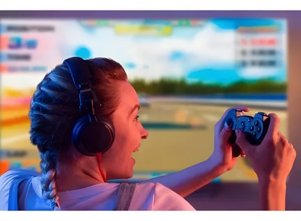 Närbild från höger sida av en person med flätat hår som bär svarta over-ear-hörlurar i plast och håller en mörkgrå spelkontroll i plast, suddig bakgrund med färgglad, lysande skärm