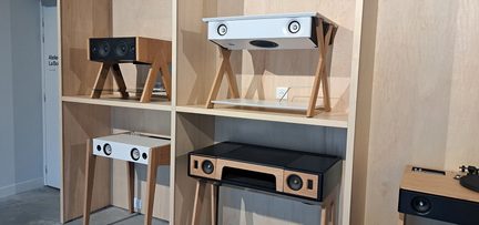 Vedere frontală a unui stand din placaj deschis la culoare, pe care sunt expuse mai multe piese de mobilier audio dreptunghiulare din lemn deschis, cu fațade albe sau negre, difuzoare circulare integrate și picioare înclinate din lemn