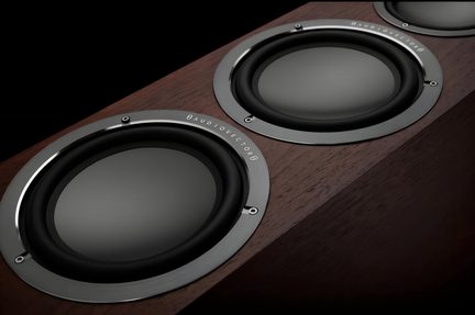 Gros plan en vue oblique d’une enceinte en bois brun foncé, trois haut-parleurs circulaires noirs cerclés d’aluminium brossé à vis apparentes