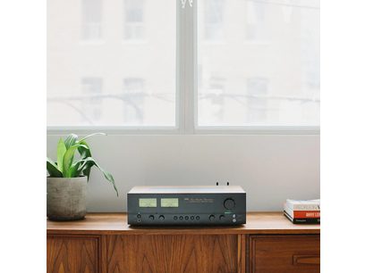 Στερεοφωνικός ενισχυτής hi-fi NAD C3050 Standard Edition Μαύρο, μπροστινή όψη, τοποθετημένος σε ξύλινο έπιπλο, με μια πράσινη φυτεία στα αριστερά