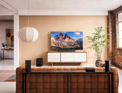 Salon moderne vu de face, canapé capitonné en cuir brun au premier plan, deux enceintes colonnes noires et un boîtier audio sur la console arrière, meuble bas blanc bordé de bois supportant un téléviseur plat affichant un paysage, suspension textile crème et plante verte en pot à droite